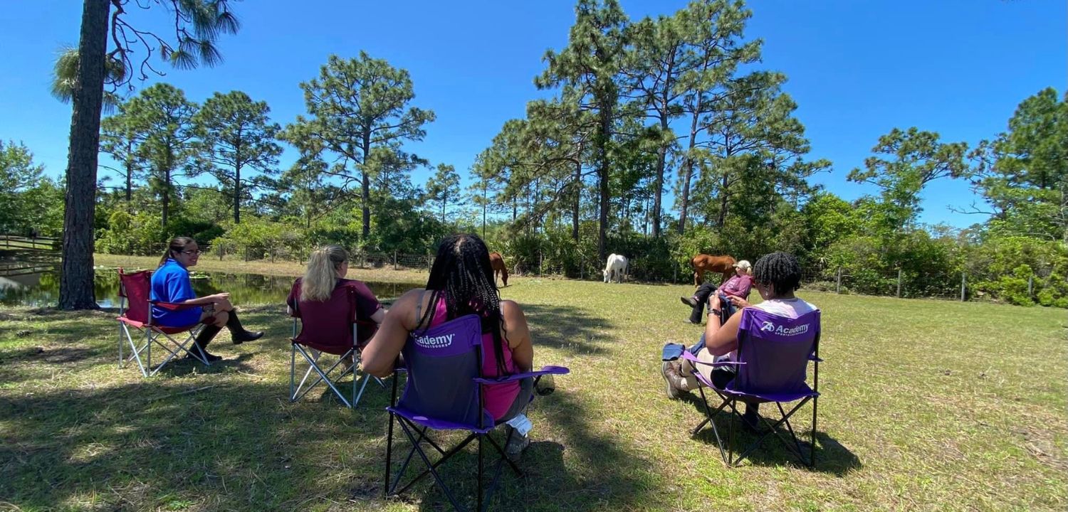 The HERD Institute_ Group Photo. ladies sitting outside in a circle with horses in a pasture.
